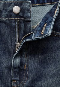 Jean en denim bleu foncé non zippé montrant une fermeture éclair métallique, un bouton et des coutures détaillées sur le tissu.