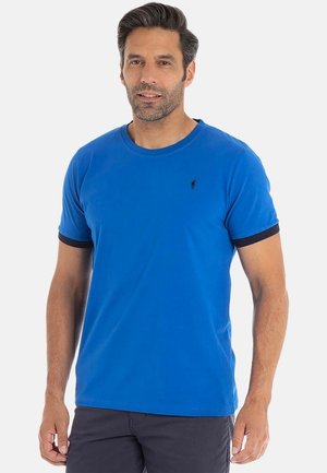 Homme portant un t-shirt bleu à manches courtes avec des bordures noires sur les manches, se tenant contre un fond blanc uni.