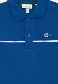 Zils polo krekls ar teksturētu audumu. Iezīmē trīs zilus pogas, baltu horizontālu svītru un zaļu apkakli. Izšūta Lacoste logo.