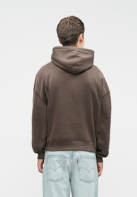 Felpa con cappuccio marrone e vestibilità rilassata, orlo e polsini a coste. Abbinata a jeans in denim azzurro, con dettagli di cuciture e tasche.