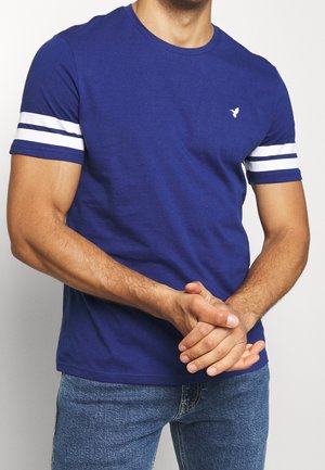 Mann trägt ein blaues T-Shirt mit weißen Doppelstreifen an den Ärmeln und einem kleinen weißen Logo auf der Brust, kombiniert mit blauen Jeans, Hände verschränkt.