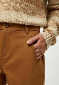 Pantaloni marroni su misura realizzati in tessuto liscio, con una tasca laterale e chiusura con bottone. Abbinati a un maglione lavorato a maglia beige e marrone a trama.