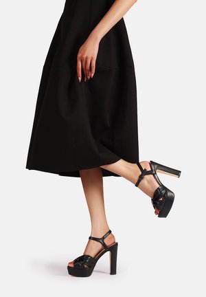 Vrouw die zwarte sandalen met hoge plateauzolen en enkelbandjes draagt en een zwarte midirok, poseren met één been omhoog.
