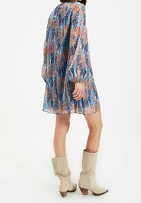 Mehrfarbiges Blumenkleid mit langen Ärmeln, transparentem Stoff, gerafftem Rücken und einer halbfigurnahen Silhouette. Kombiniert mit beigen Stiefeletten mit Absatz.