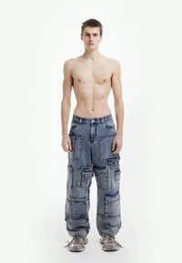 Junger Mann, der aufrecht steht, oberkörperfrei, trägt lockere blaue Cargo-Jeans mit mehreren Taschen und grauen Sneakern, vor einem einfarbigen weißen Hintergrund.