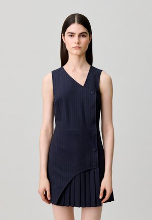 Giovane donna con lunghi capelli scuri indossa un abito senza maniche blu navy a righe sottili con davanti asimmetrico abbottonato e pannello di gonna plissettata.