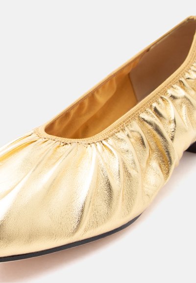 Χρυσό μεταλλικό μαλακό δερμάτινο παπούτσι με επίπεδη σόλα, με ελαστική επάνω άκρη και στρογγυλεμένη μύτη, εμφανίζεται σε κοντινό πλάνο σε λευκό φόντο.