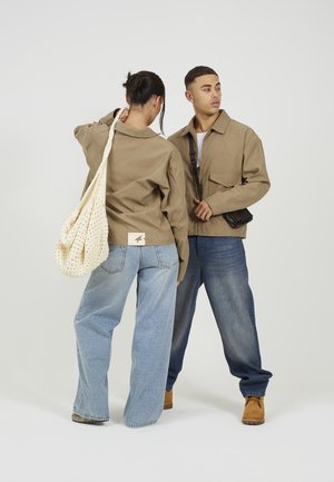 Dwie osoby w beżowych kurtkach i luźnych dżinsach; jedna niesie kremową dzianą torbę na ramię, druga nosi beżowe botki i czarną torbę przewieszoną przez ciało.