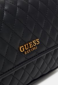 Sac à main en cuir noir matelassé avec un logo "GUESS" doré accompagné de la mention "EST 1981". Texture lisse avec motif en losange et coutures robustes.