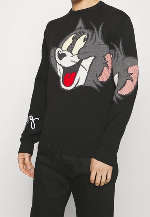 Czarny sweter z dużym nadrukiem przedstawiającym twarz Toma, kota z bajki Tom i Jerry, noszony przez osobę w czarnych spodniach na tle o jednolitym kolorze.