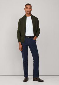 Donkergroene zip-up sweater over een wit T-shirt, marineblauwe broek en bruine schoenen. Eenvoudig ontwerp met een slim-fit pasvorm en een gladde textuur.