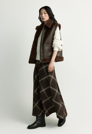 Woman wearing a brown fur-trimmed leather vest, white sweater, long plaid skirt, and black boots standing against a plain background.