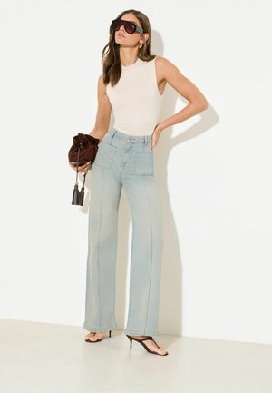 Žena v bielom tielku bez rukávov, svetlomodrých širokých džínsach s vysokým pásom, čiernych sandáloch na opätku, s veľkými slnečnými okuliarmi a držiaca hnedú kabelku s strapcami.