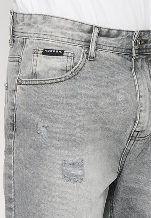 Šedé džíny z denimu s opotřebovaným designem a třepenými záplatami. Detailní švy a logo na pásku. Klasický pětikusový styl.