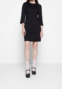 Musta shift-mekko, jossa kolme neljäsosahihnaa ja vyötäröllä kaksi kangaspäällysteistä nappia. Yhdistetty vaaleanpunaisten nilkkasukien ja mustien puukengien kanssa.