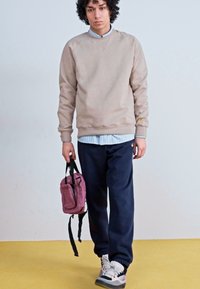Beige Sweatshirt mit gerippten Bündchen, marineblaue Hose und einer kleinen pinken Tasche. Das Model trägt ein gestreiftes Hemd mit Kragen darunter.