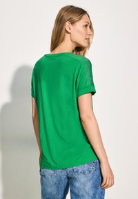Grøn kortærmet t-shirt lavet af blødt stof med rulleærmer; har en glat tekstur og en afslappet pasform. Sæt af lyseblå jeans vist.