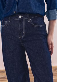 Ciemnoniebieskie dżinsy z dopasowanym stanem, prostymi nogawkami i kontrastującym jasnym szwem. Posiadają przednie kieszenie i zapięcie na metalowy guzik.