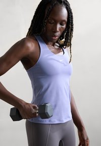 Kvinde i en lavendel farvet tanktop holder en sort hexagonal håndvægt og ser ned på den, mod en lys ensfarvet baggrund.
