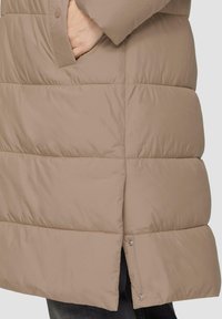 Puffermantel in hellbeige mit gestepptem Design, seitlichen Taschen und Druckknopfverschluss am Saum, mit einer glatten, glänzenden Textur.