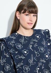 Blusa blu navy con stampa floreale, dettagli arricciati sulle spalle, scollatura rotonda e chiusura con bottoni. Realizzata in tessuto leggero con una texture liscia.