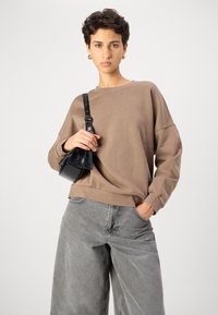 Sudadera marrón de corte relajado, con puños acanalados y escote redondo, combinada con vaqueros de pierna ancha grises y un pequeño bolso negro.