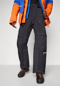 Czarne spodnie outdoorowe o luźnym kroju, z zamykanymi na zamek kieszeniami, wzmocnionymi szwami oraz logo "Mammut" na dolnej części nogawki.