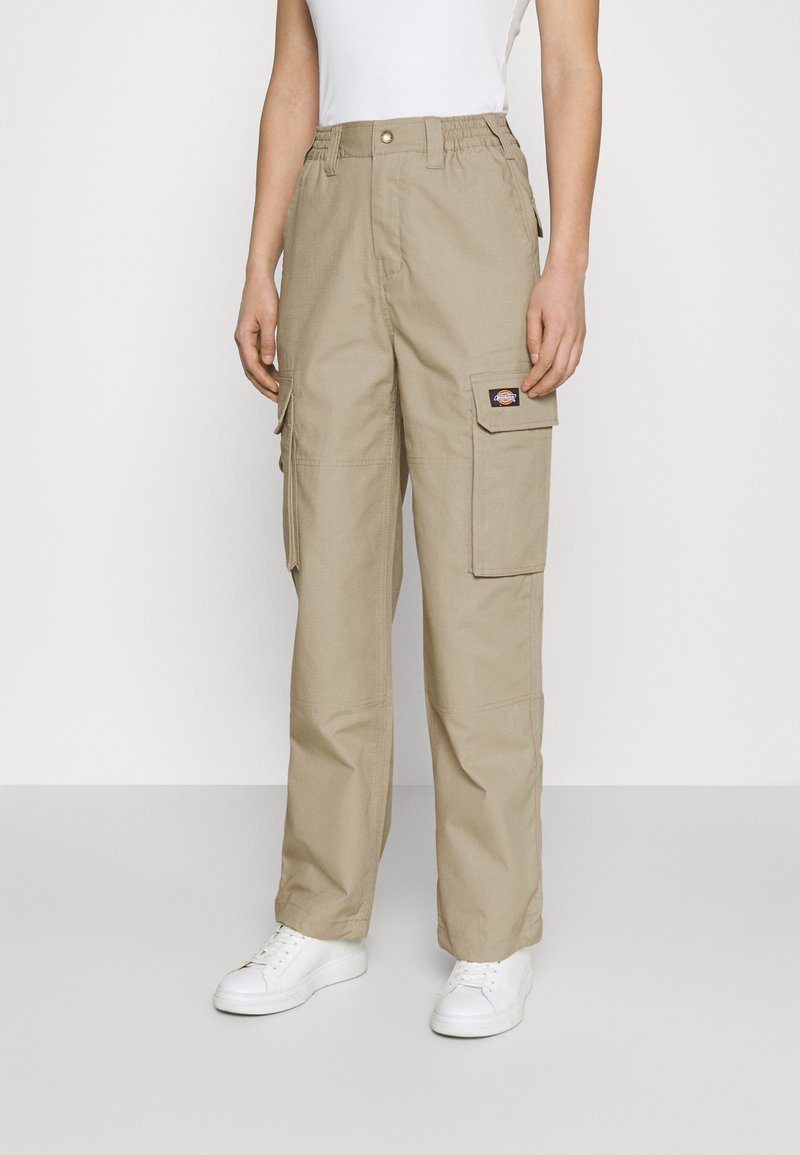 Beige cargobroek met elastische tailleband, meerdere zakken en een lichte glans. Gecombineerd met witte sneakers en een aansluitend wit topje.