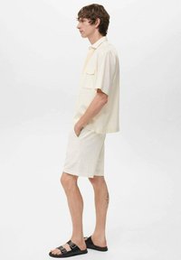 Chemise légère crème à manches courtes avec poche, associée à un short blanc texturé et des sandales noires à boucles. Design épuré et minimaliste.