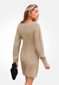 Strickkleid in Beige mit langen Zopfmuster-Ärmeln, einer taillierten Silhouette und einem weiten Ausschnitt. Das Model hält eine schwarze Clutch.