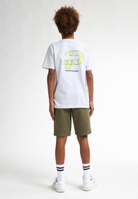 Witte T-shirt met korte mouwen en een neon groene print op de achterkant, gecombineerd met olijfgroene shorts en witte sneakers. Zwart-witte gestreepte sokken.