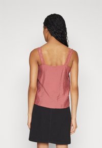 Vaaleanpunainen toppi, jossa on pitsiset olkaimet, pehmeä rakenne ja hieman valuva muotoilu. Yhdistetty mustaan hameeseen, joka näyttää rentoa istuvuutta ja epämuodollista tyyliä.