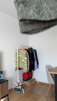 Clothing rack with various sweaters and shirts. A green striped sweater is prominent. A red sports bag and potted flower decorate the space.