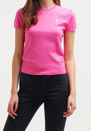 Woman wearing bright pink short-sleeve fitted knit top with small white logo on left chest and black pants against a plain background.
