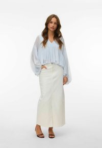 Femme debout devant un fond blanc portant un chemisier transparent bleu pâle à manches bouffantes, une longue jupe blanche et des sandales ouvertes.