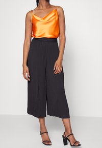 Frau trägt ein ärmelloses Oberteil aus orangefarbenem Satin, schwarze weitgeschnittene cropped Hosen, schwarze Absatzsandalen und hält eine kleine Tasche mit Kettenriemen.