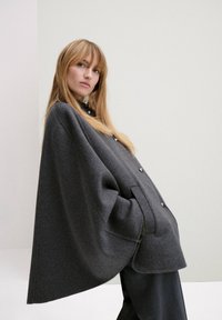 Cape en laine grise avec une silhouette décontractée, dotée de grands boutons, de poches latérales et d'une finition texturée. L'arrière-plan est minimaliste.