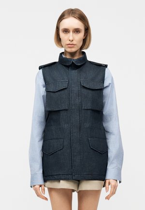 Νέα γυναίκα με ανοιχτό μπλε ριγέ πουκάμισο και αμάνικο σκουρομπλε τζιν γιλέκο με πλαϊνές τσέπες, στέκεται μπροστά σε απλό φωτεινό φόντο.