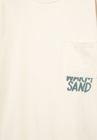 Lys beige bomulds T-shirt med en lomme på venstre bryst og blå tekst, der læser "WARM SAND" i en fed, afslappet skrifttype.