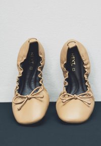 Beige nahkainen ballerina-sandaalit, joissa on pyöreä varvas, rypytetty aukko ja nauhakoriste edessä. Musta sisäosa kultaisella brändäyksellä.
