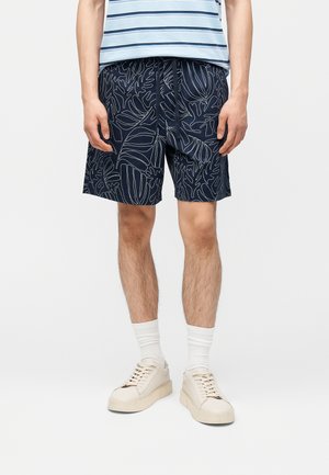 Férfi sötétkék rövidnadrágban, fehér levélmintákkal, fehér tornacipőben, fehér zokniban és kék csíkos ingben, egyszínű háttér előtt állva.