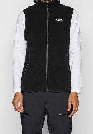 Sort fleeced vest uden ærmer med lynlås og The North Face-logo, båret over en langærmet hvid skjorte og sorte bukser med lynlåslommer.