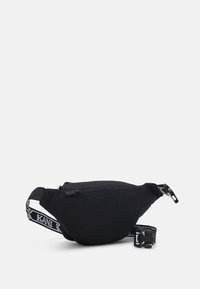 Musta kangaskotelo vetoketjukiinnityksellä, kaareva muoto, säädettävä hihna, joka esittelee kontrastivärisen logon, sekä sileä pinta.