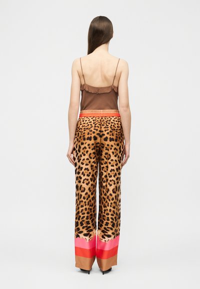 Γυναίκα με μακριά καστανά μαλλιά, φορώντας καφέ τοπ και παντελόνι φαρδύ με πλάγιες ρίγες λεοπάρ σε ροζ και κόκκινους χρωματικούς τόνους, εμφανιζόμενη από πίσω.