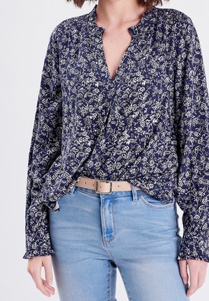 Femme portant un chemisier à fleurs bleu marine à manches longues rentré dans un jean bleu clair avec une ceinture beige sur un fond uni.