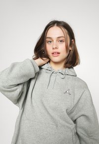 Grå hettegenser med snøring i halsen, stor frontlomme og svart Jumpman-logo på brystet. Myk stoffkvalitet.