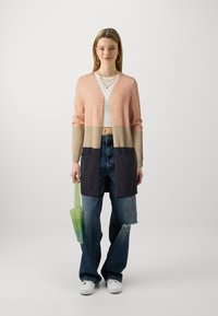 Cardigã longo em rosa pastel, bege e azul marinho, com dois bolsos frontais. Usado sobre uma blusa branca e combinado com jeans azuis desfiados. Bolsa verde.