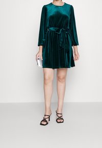 Teal-värinen samettinen mekko, jossa on pitkät hihat ja vyötärösidos. Mekossa on pyöreä kaula-aukko. Se on yhdistetty mustiin remmikenkiin ja hopeiseen iltalaukkuun.