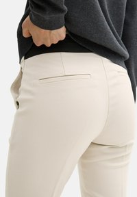 Elfenbeinfarbene, maßgeschneiderte Hose mit glatter Textur, mit einer Gesäßtasche und einer figurbetonten Silhouette. Kombiniert mit einem dunkelgrauen Pullover.