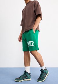 Brown oversized t-shirt, green Nike shorts with white logo, and black/teal high-top sneakers with white soles, worn with white crew socks.